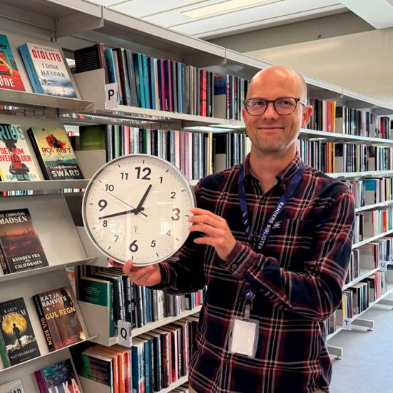 Mads Einar Nielsen holder et væg-ur i biblioteksrummet.