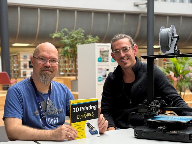 To mænd sidder ved et bord med en bog og en 3D-printer
