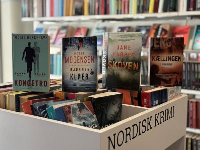 Fire bøger stillet på højkant på en krybbe med bøger med bogreoler i baggrunden