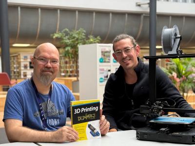 To mænd sidder ved et bord med en bog og en 3D-printer